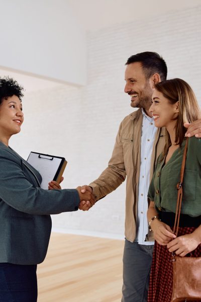 Happy couple and their real estate agent shaking hands in a new house. Happy real estate agent greeting a couple during open house day.